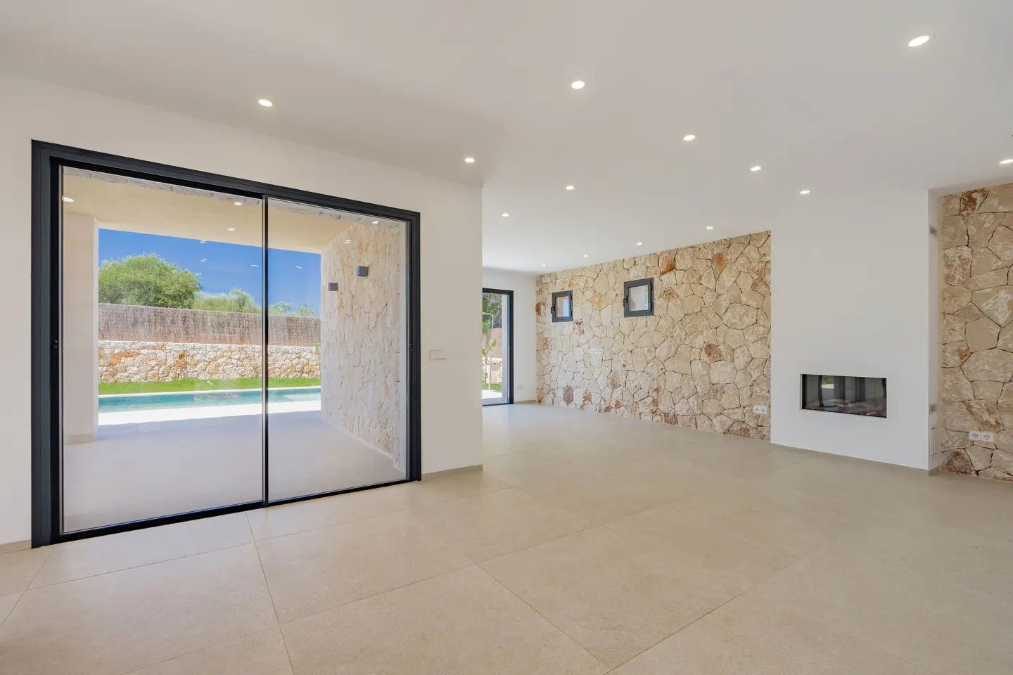 Bright, empty room with beige tile floor and stone accent wall. Black-framed sliding glass doors open to a pool and patio.