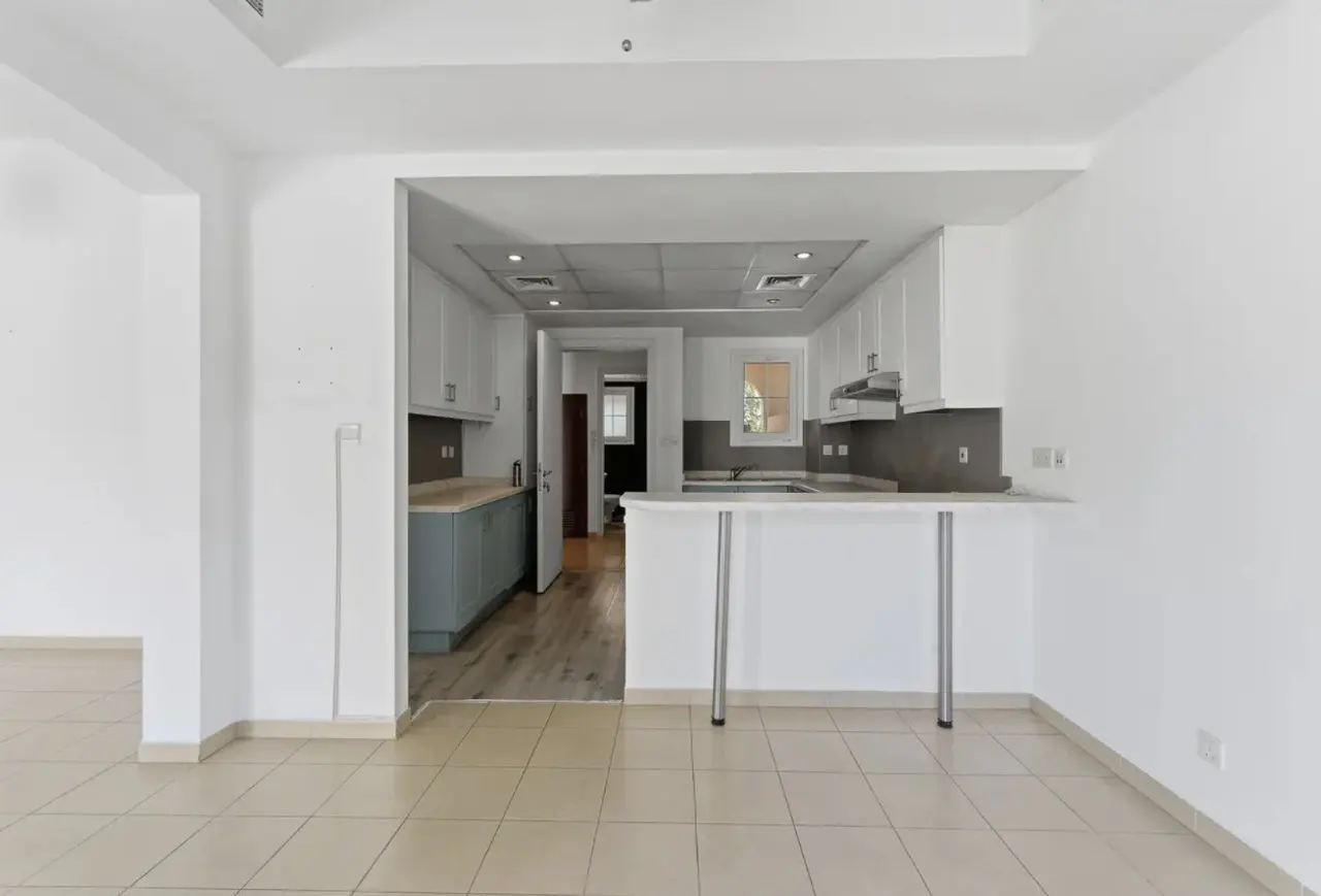 A bright, empty room with beige tile floors leads to a kitchen with white cabinets and a breakfast bar.