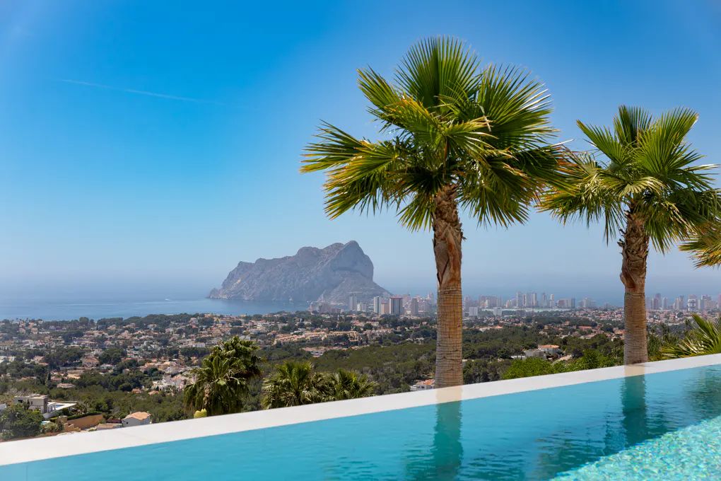 Infinity pool with palm trees overlooking a coastal city and the Penon de Ifach rock formation under a clear blue sky.