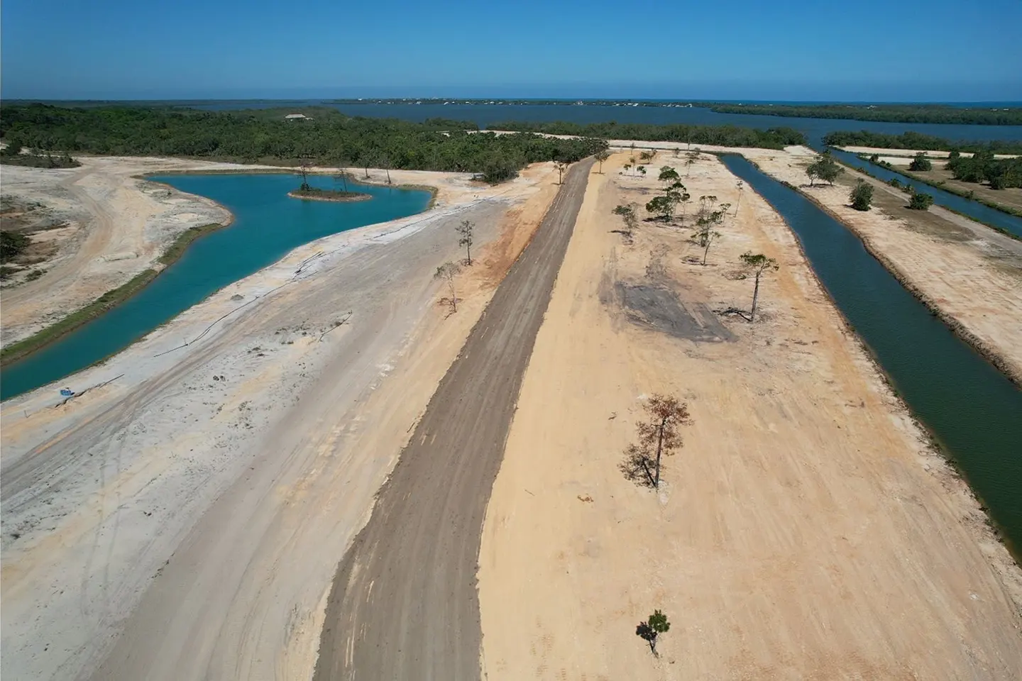 Aerial view of a real estate development with canals, a road, and turquoise water features. Trees dot the sandy landscape under a clear blue sky.