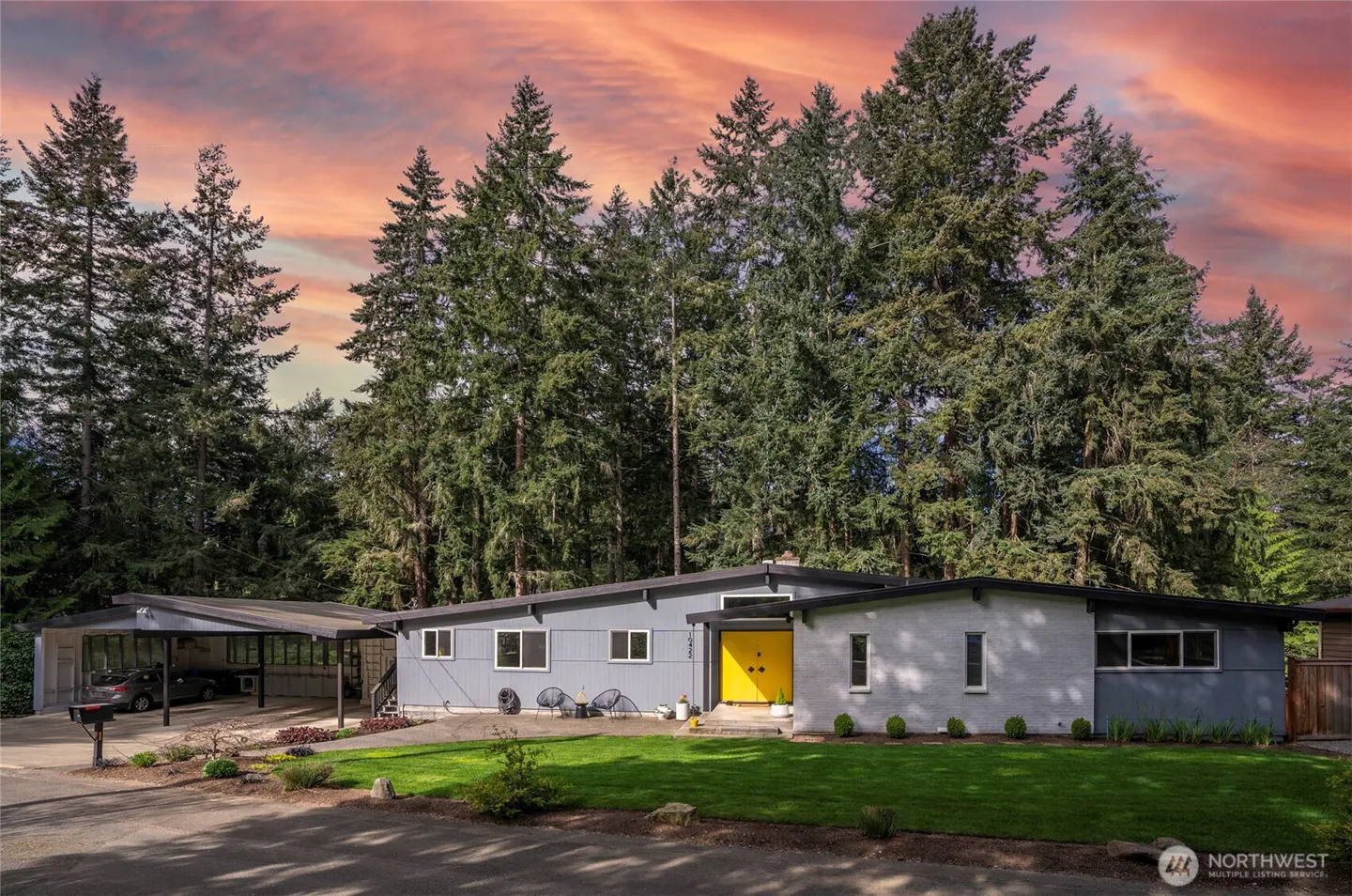 Gray mid-century modern home with a bright yellow front door and a carport, set against a backdrop of tall green trees and a colorful sunset.