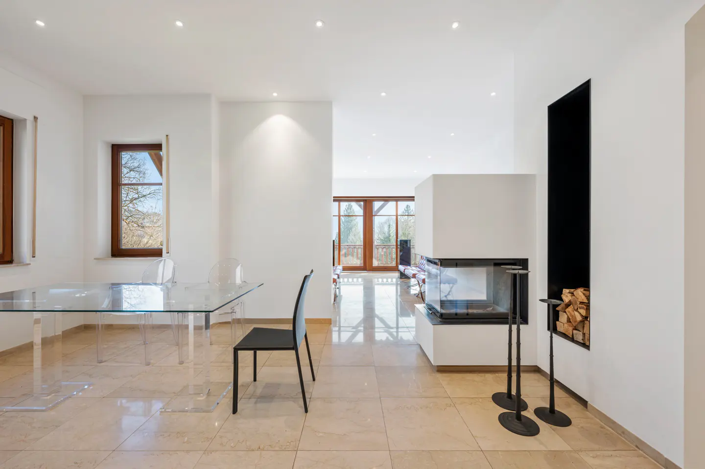 Bright, modern room with a glass table, black chair, and fireplace with wood storage. Windows offer a view of trees.