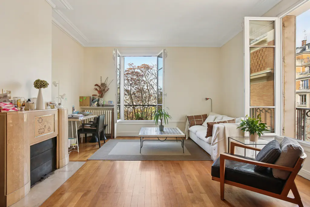 Bright living room with hardwood floors, fireplace, desk, and white sofa. Open windows reveal trees and city views.