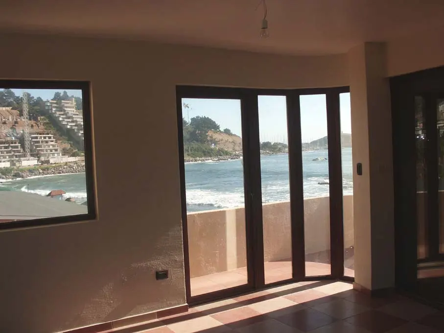 Interior view of a room with a window and sliding glass doors overlooking the ocean. The walls are beige and the floor is tiled.