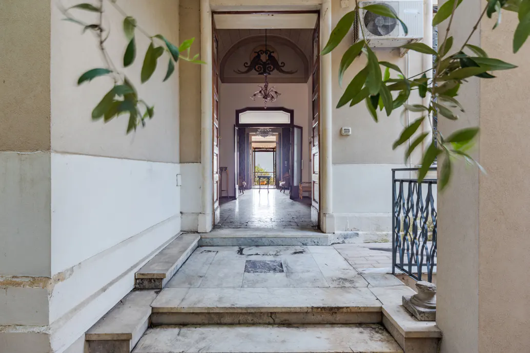 View of a hallway with marble floors and white walls, leading to an open doorway with a view of greenery.