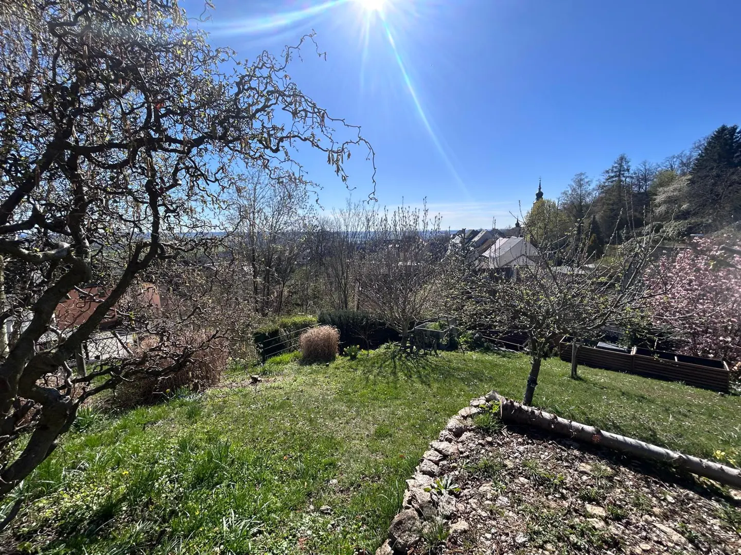 Sunny view of a green lawn with trees, a stone wall, and a town in the distance under a blue sky.