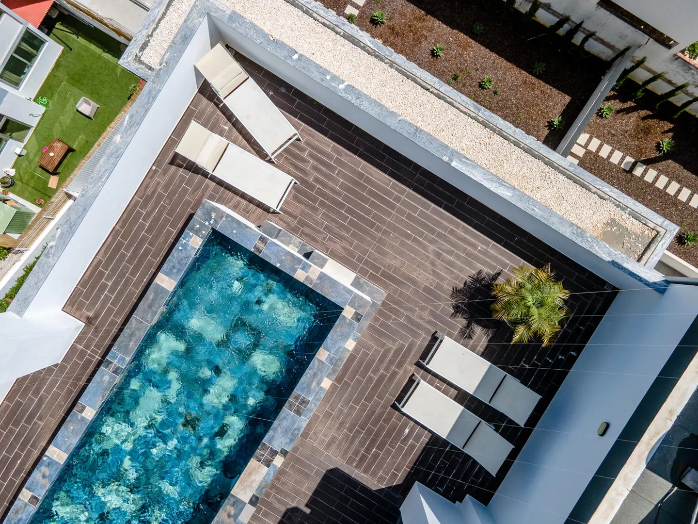 Aerial view of a rooftop pool with blue water, surrounded by lounge chairs and brown brick flooring. A small palm tree sits nearby.