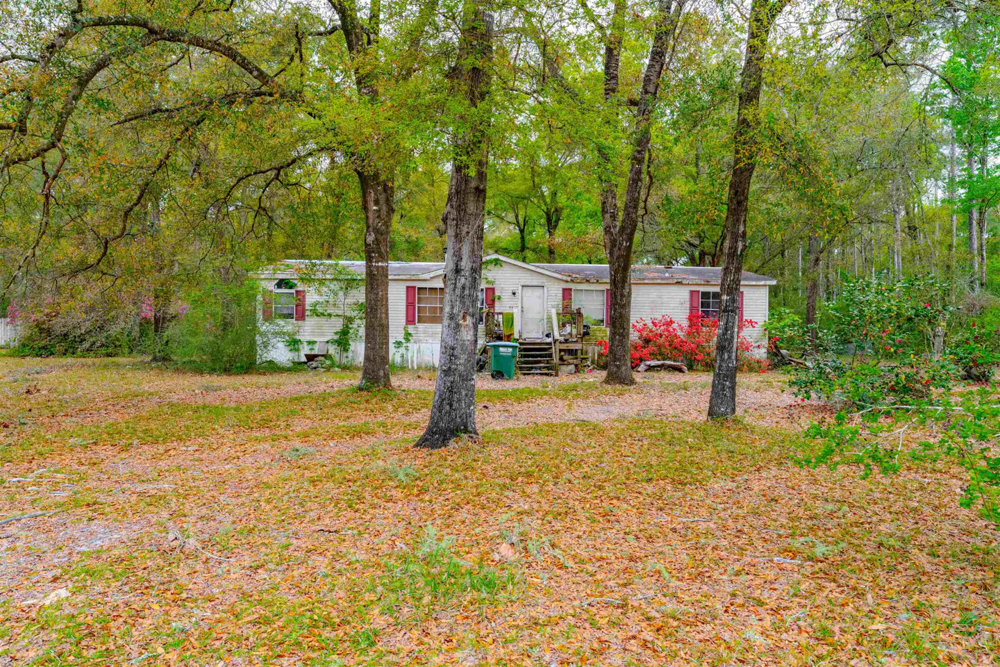 Exterior view of a white single-story home with red shutters, surrounded by trees and a yard covered in fallen leaves.