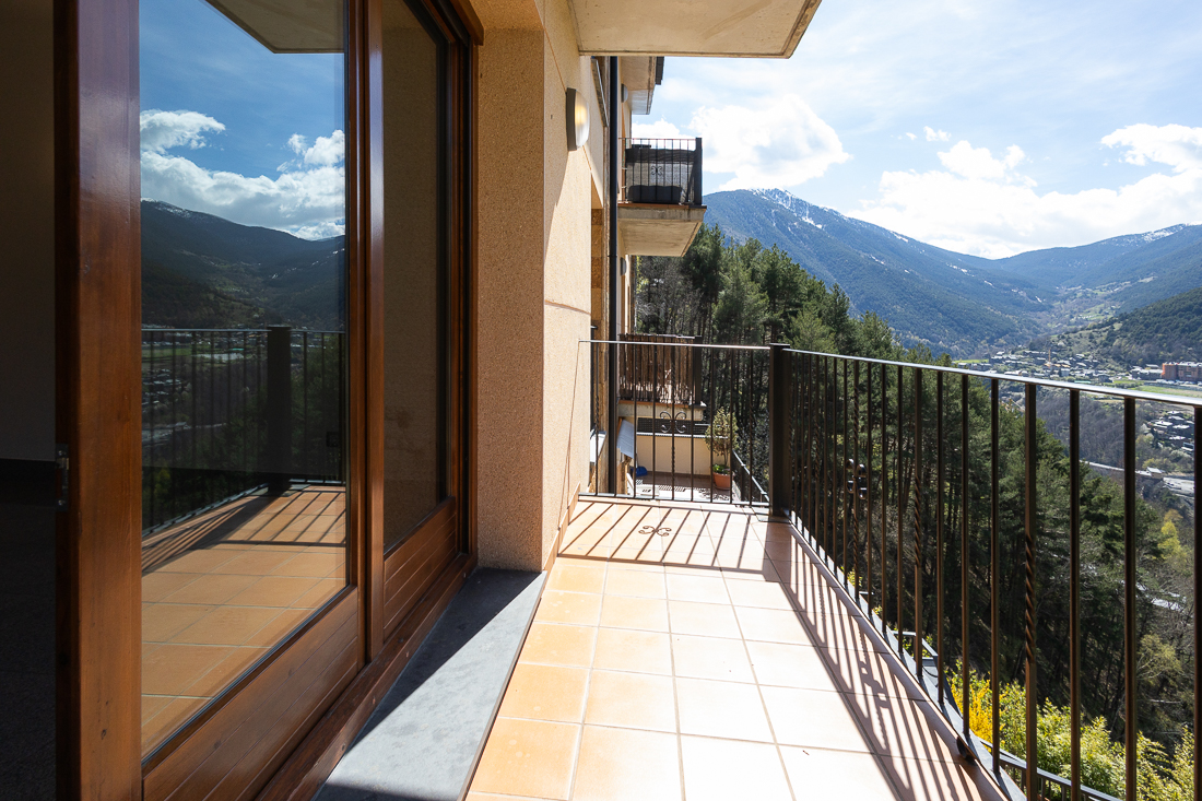 Balcony with brown tile floor and black railing overlooks a mountain view with green trees and a town in the distance.