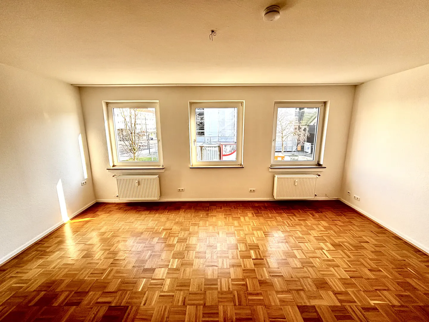 Empty room with parquet floor, white walls, and three windows. Radiators are under each window. A smoke detector is on the ceiling.