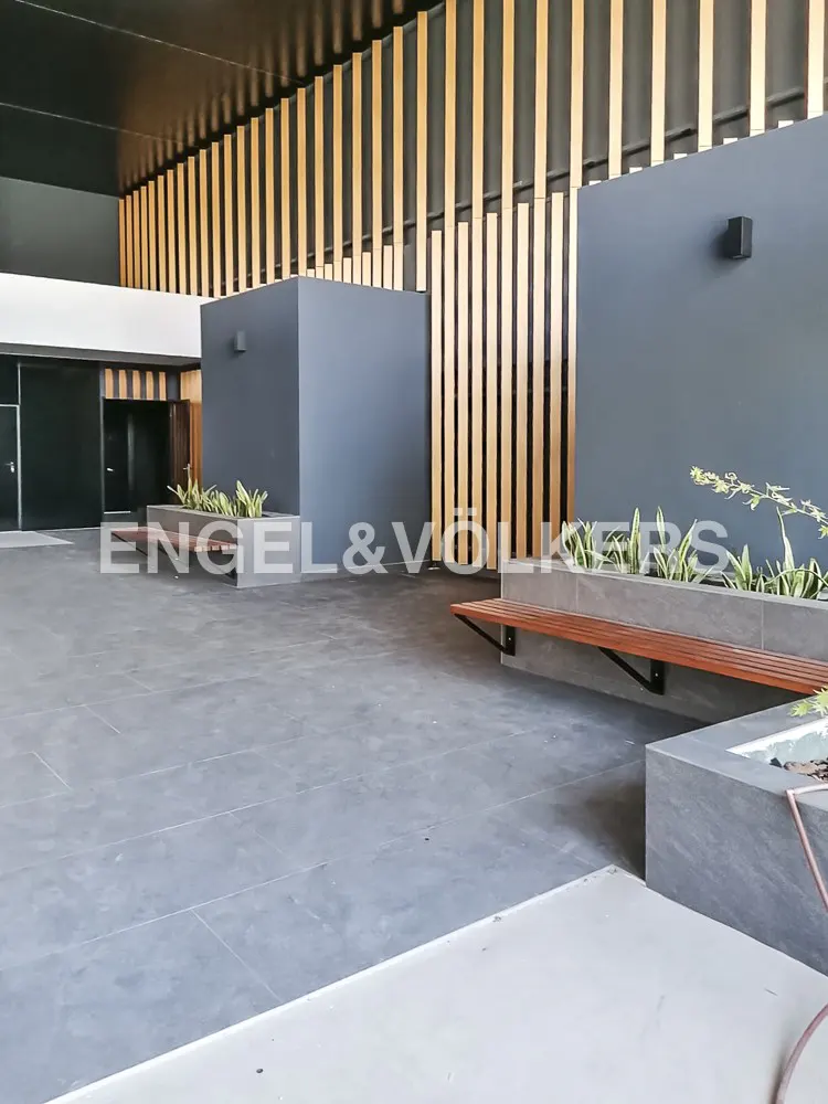 Modern lobby with gray tile floor, wood benches, and planters. Gray walls are accented by vertical wood slats. Engel & Völkers logo visible.
