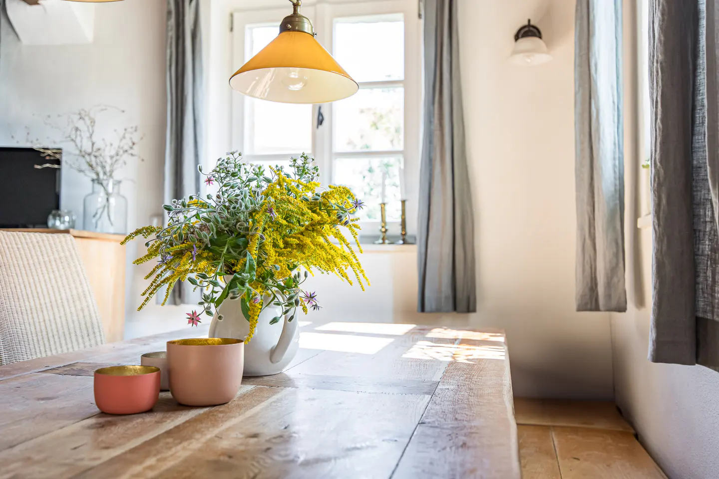 A bright room features a wooden table with a vase of yellow flowers, pink cups, and natural light from a window with gray curtains.