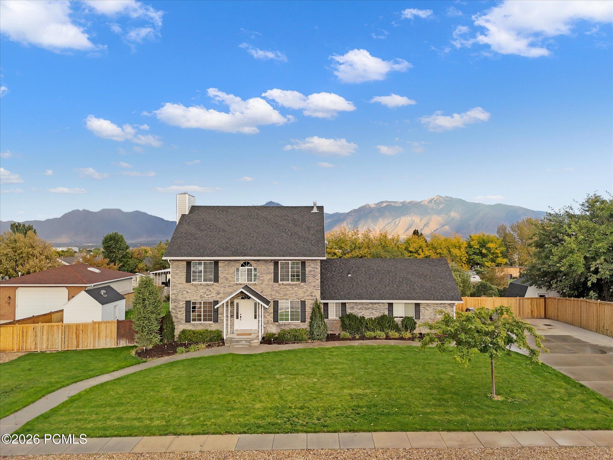 Two-story brick home with a dark shingled roof, large front lawn, and scenic mountain views under a blue sky. [AI-generated text]