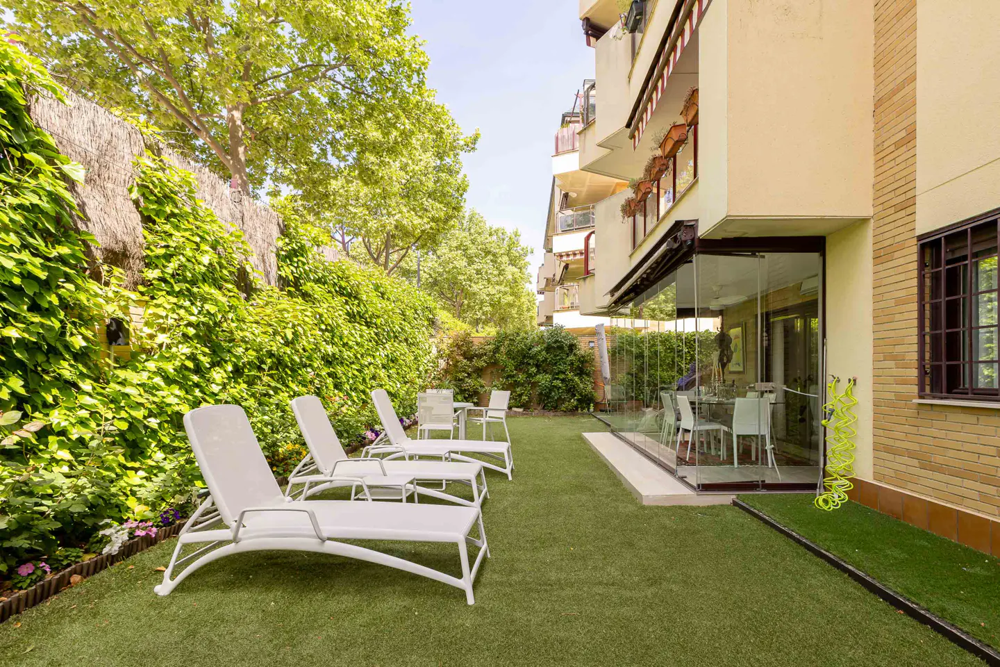 A backyard with white lounge chairs on green grass, next to a building with a glass-enclosed patio.