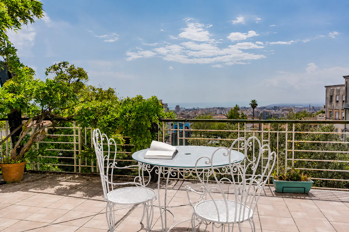 Balcony view with white metal table and chairs, book, and city skyline. Green foliage and blue sky in the background.