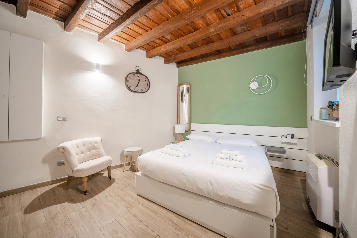 Bedroom with white bed, green accent wall, exposed wooden ceiling beams, white armchair, and a wall clock.