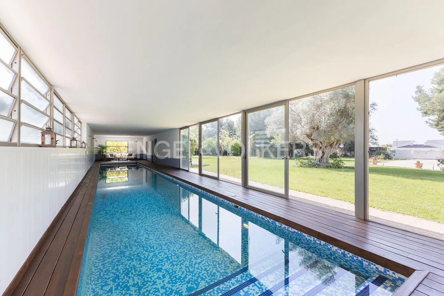 Indoor pool with blue mosaic tiles and wood deck. Large windows overlook a green lawn with trees. Lanterns line the wall.