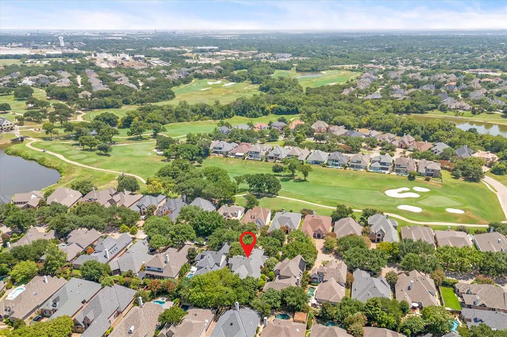 Aerial view of a residential area with houses, trees, and a golf course in the background. A red pin marks a specific property.