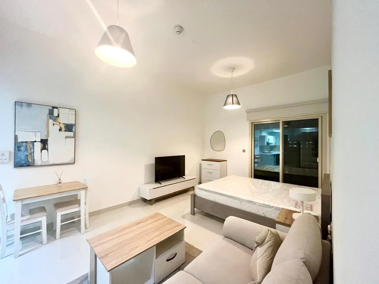 Bright studio apartment with white walls, beige sofa, bed, TV, and a small dining table. A sliding glass door leads to a balcony.