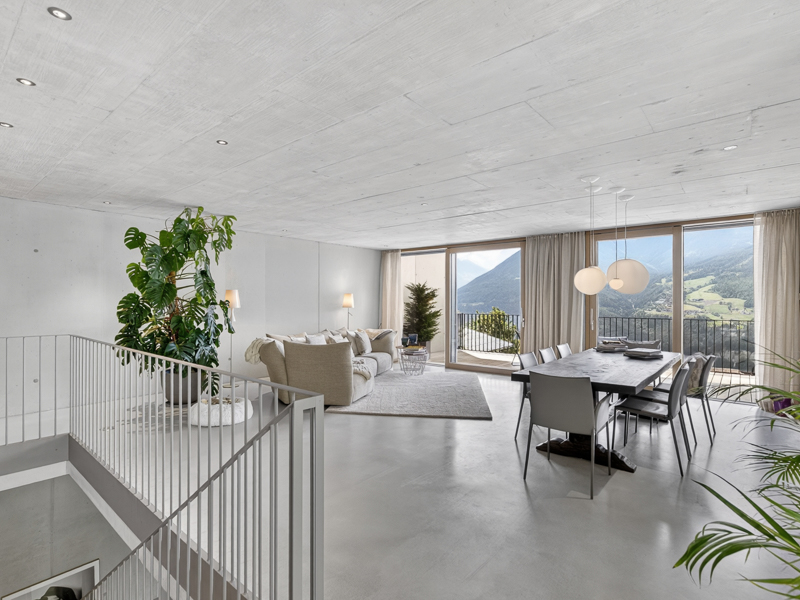 Modern open-plan living area with a beige sofa, dining table, concrete ceiling, and balcony overlooking mountain views.