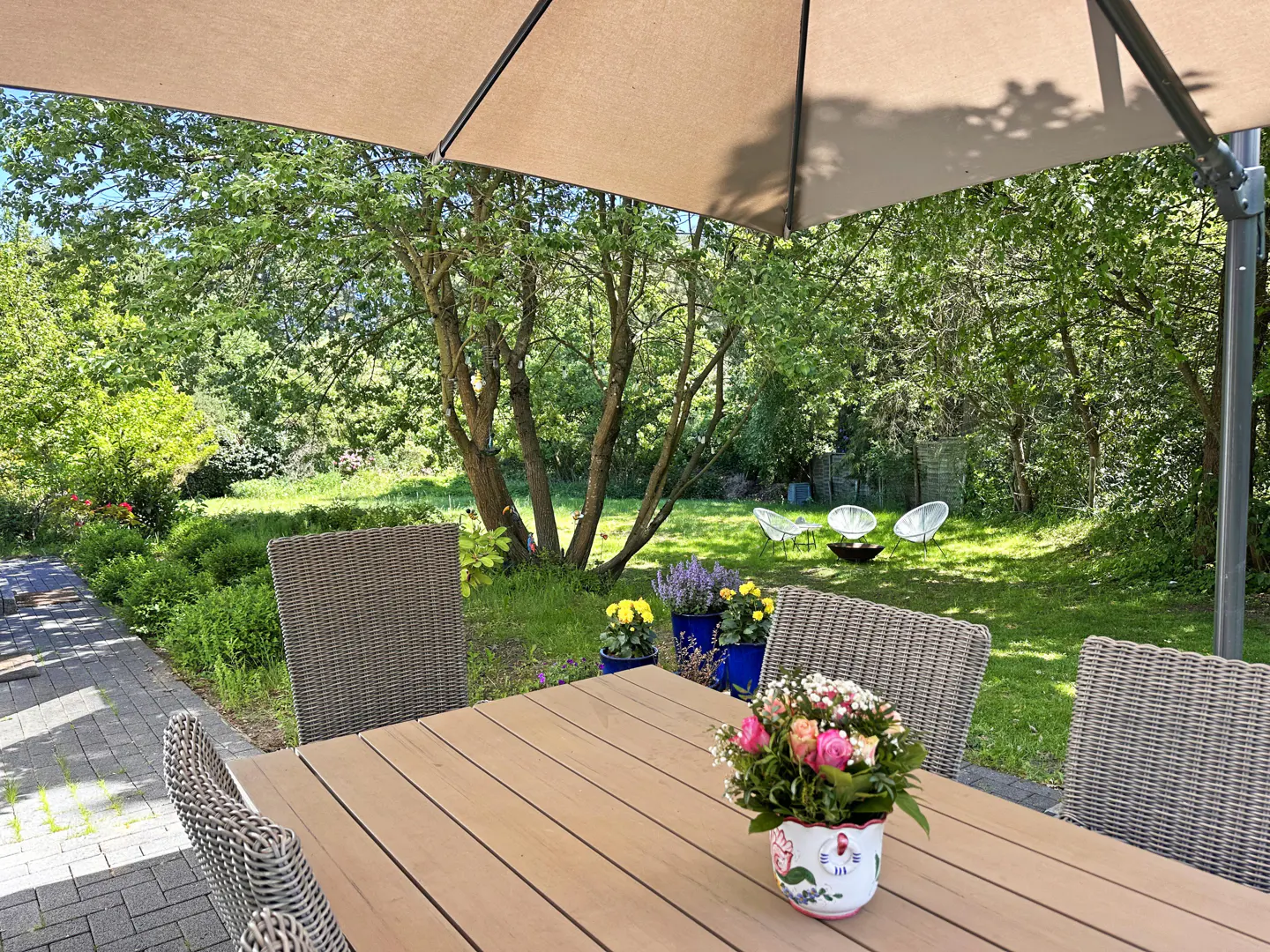Outdoor patio with a wooden table, wicker chairs, and a floral centerpiece under a large umbrella, overlooking a green yard.