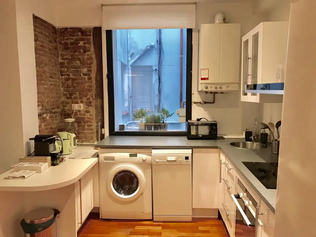 Small kitchen with white cabinets, wood floors, exposed brick wall, washing machine, dishwasher, and a window view.