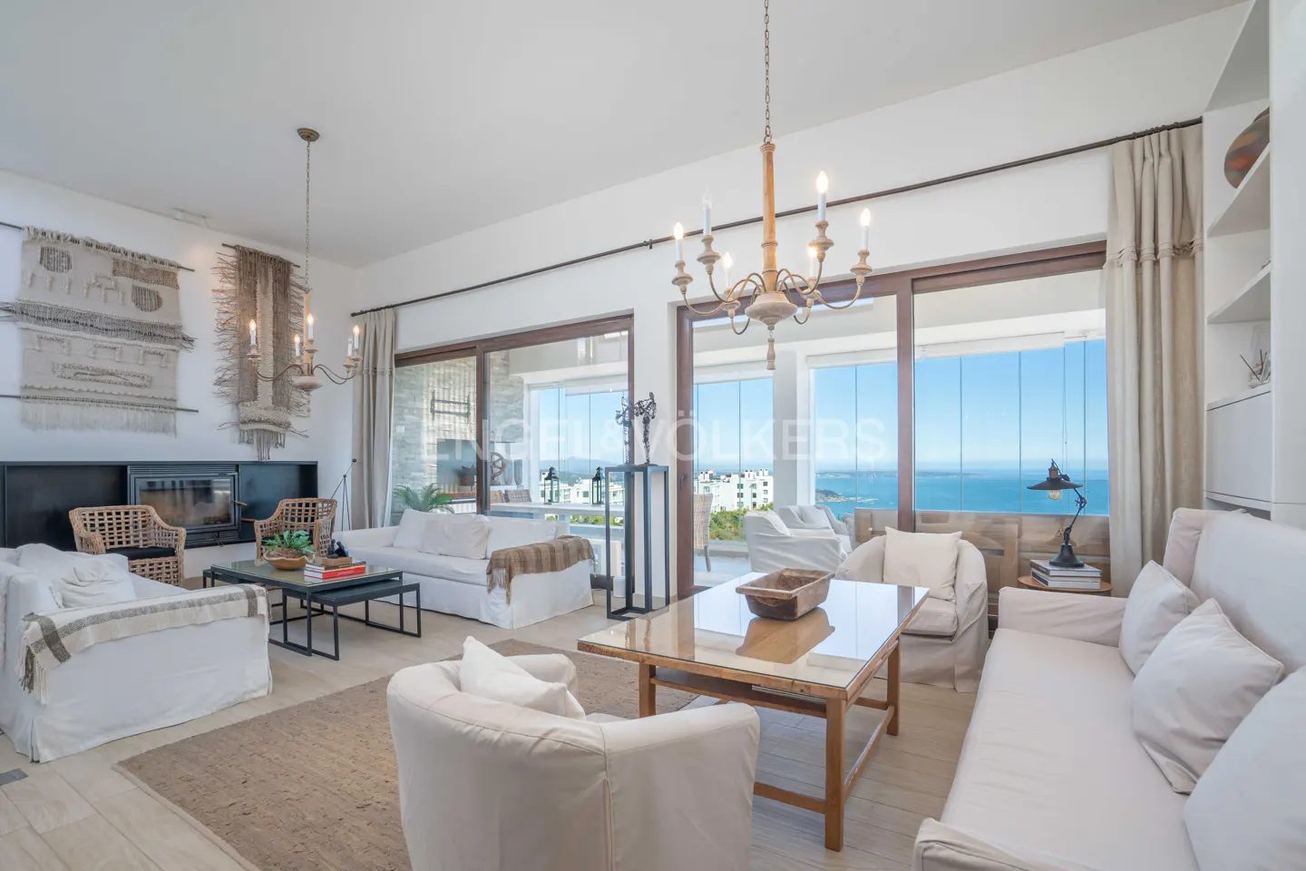 Bright living room with white sofas, a wooden table, and a chandelier. Large windows offer ocean views. A fireplace and woven wall hangings add texture.