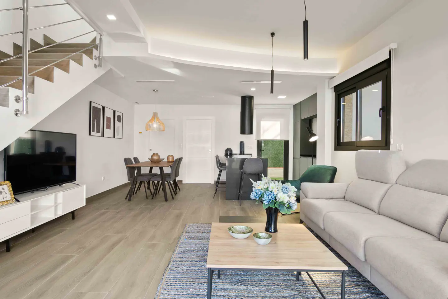 Bright, modern living space with a beige sofa, wood floors, and a staircase. A dining table and kitchen island are visible in the background.