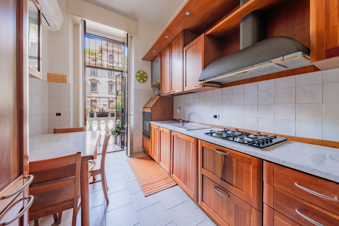 Galley kitchen with wood cabinets, white tile backsplash, marble countertops, and a balcony door overlooking city buildings.