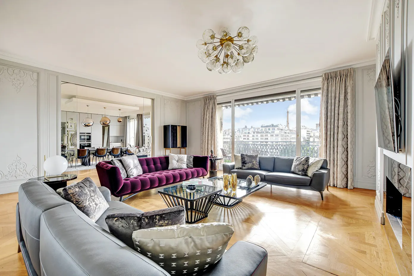 Bright living room with gray and purple sofas, glass coffee tables, and a view of Paris with the Eiffel Tower.