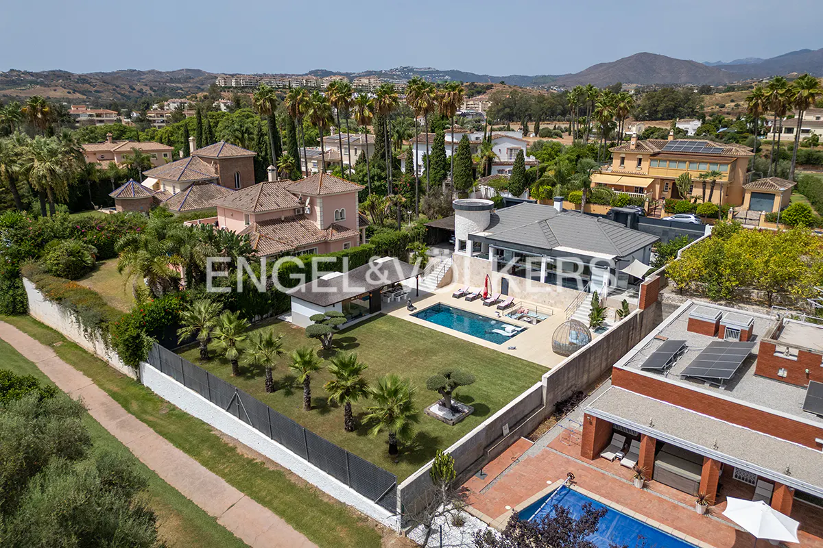 Aerial view of luxury homes with pools, palm trees, and green lawns under a blue sky. Engel & Völkers logo is visible.