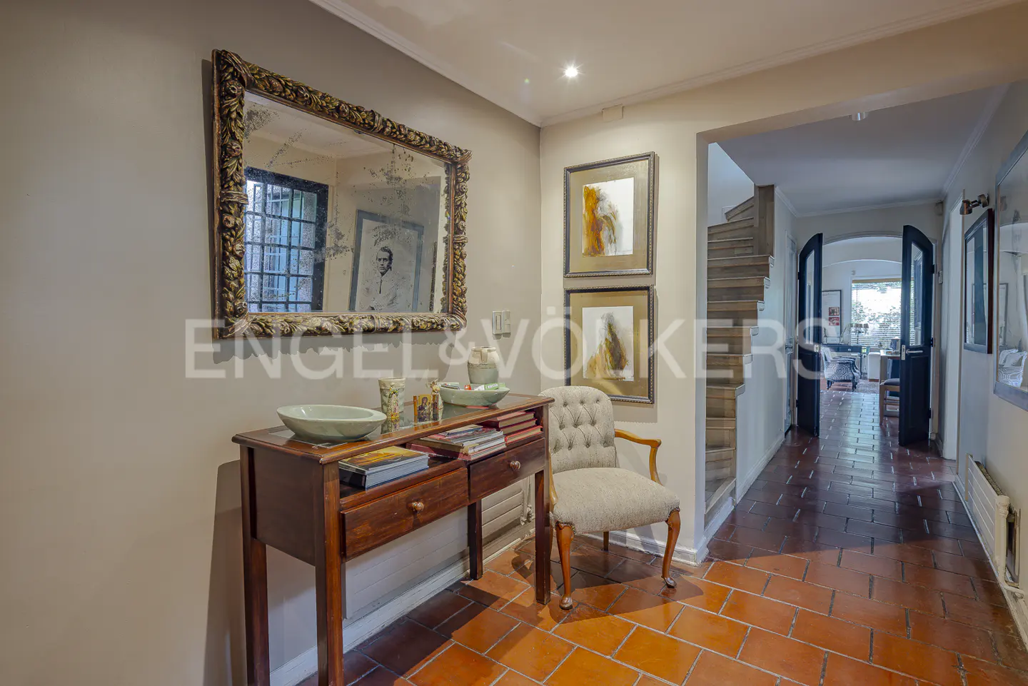 A hallway with terracotta tile floors, a wooden console table, a tufted armchair, and a large ornate mirror on the wall.