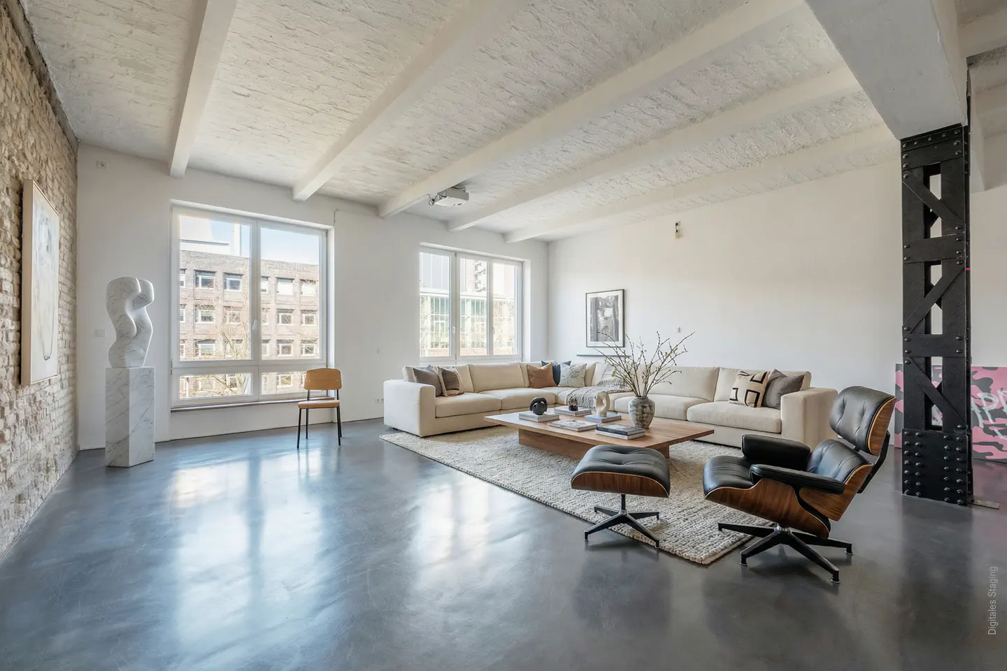 Bright, modern living room with white walls, exposed brick, and concrete floors. A large sectional sofa sits on a woven rug, with a wood coffee table and black leather chair. Large windows provide natural light.
