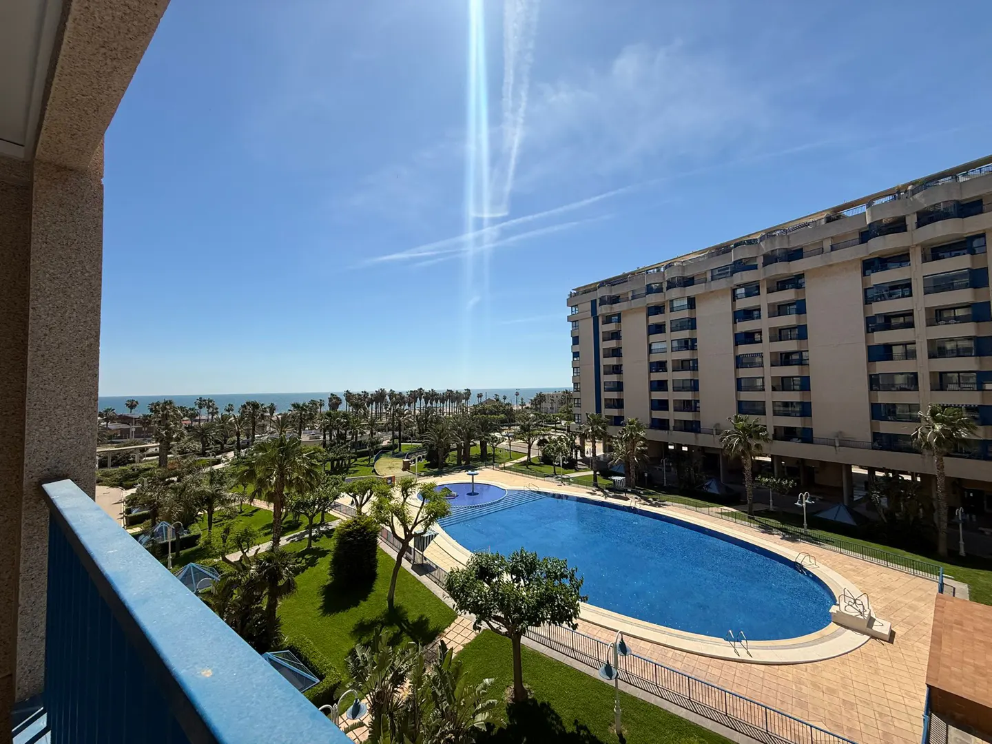 View from a balcony overlooking a blue pool, palm trees, and a building under a sunny sky.