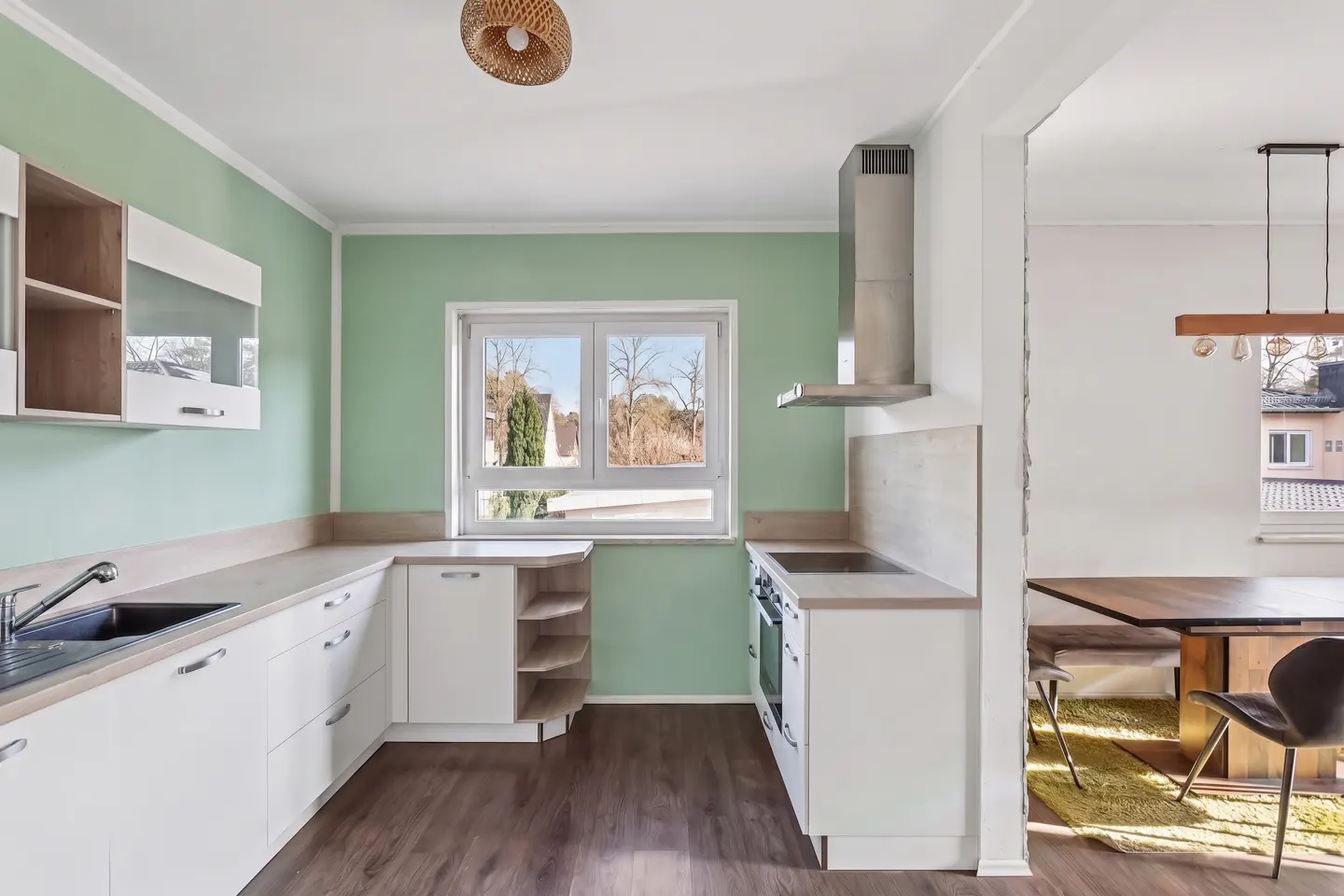 A bright kitchen with light green walls, white cabinets, and a stainless steel range hood. A window overlooks trees. A dining area is visible.