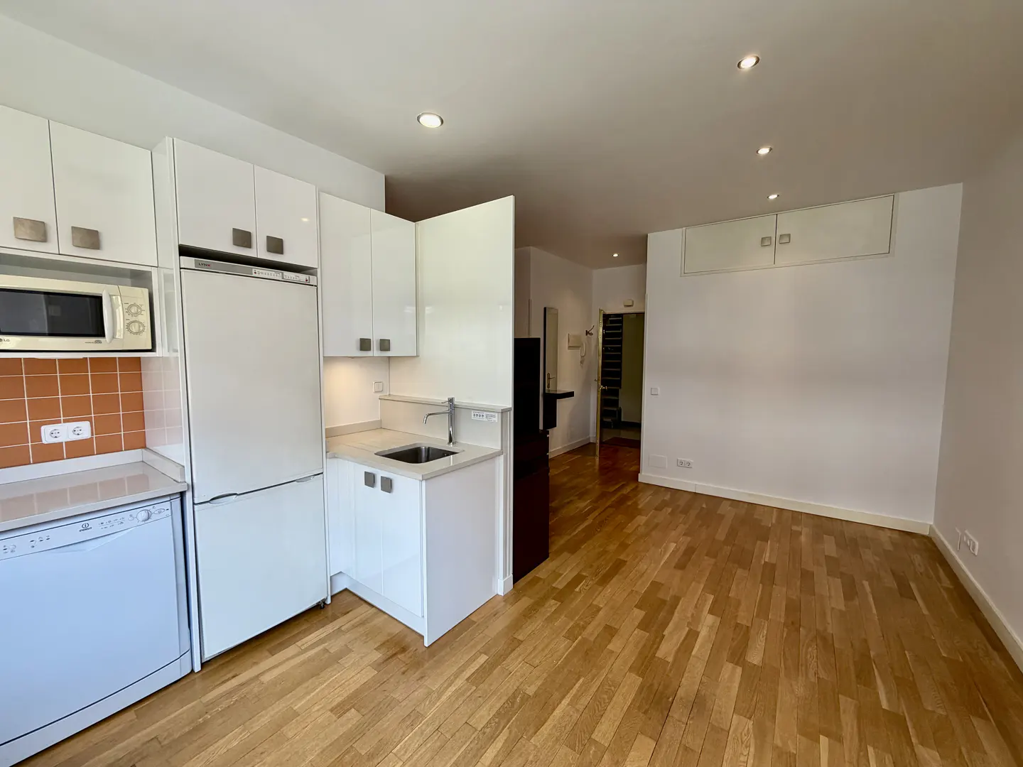 Bright studio apartment with hardwood floors, white walls, and a compact kitchen featuring white cabinets and appliances.