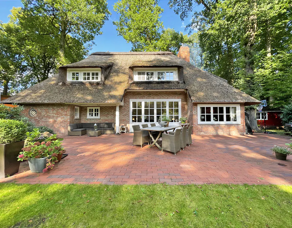 Brick house with a thatched roof, white windows, and a brick patio with outdoor furniture. Trees surround the house.