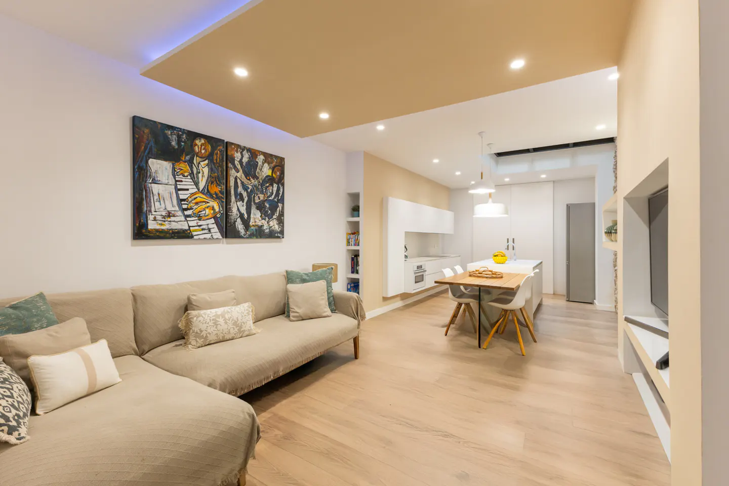 A bright, modern living space with a beige sectional sofa, abstract art, and an open kitchen with an island and dining table.