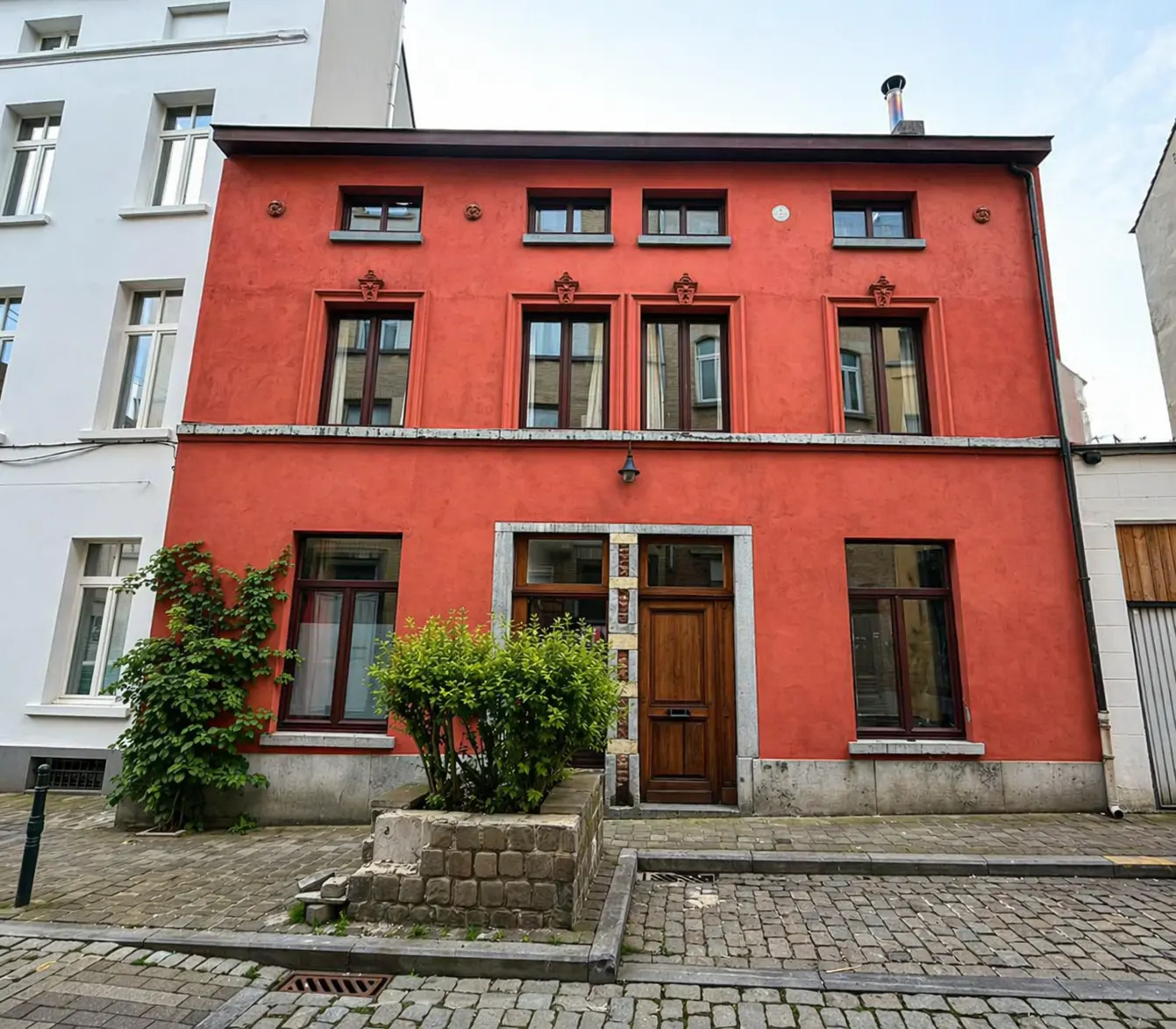Red two-story building with brown trim and a wooden door. A small bush sits in front of the building. Cobblestone street.