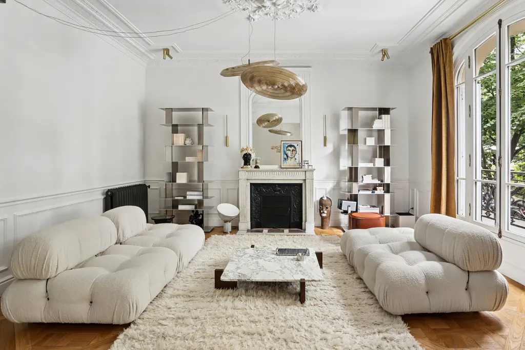Bright living room with two cream-colored modular sofas, a marble coffee table, a fireplace, and tall metal shelving units.