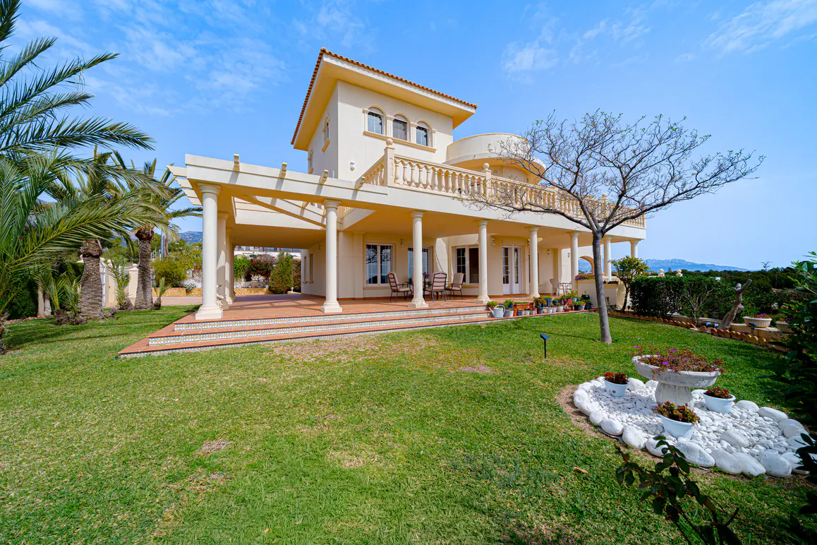 Beige two-story house with a red tile roof, columns, and a green lawn under a blue sky.