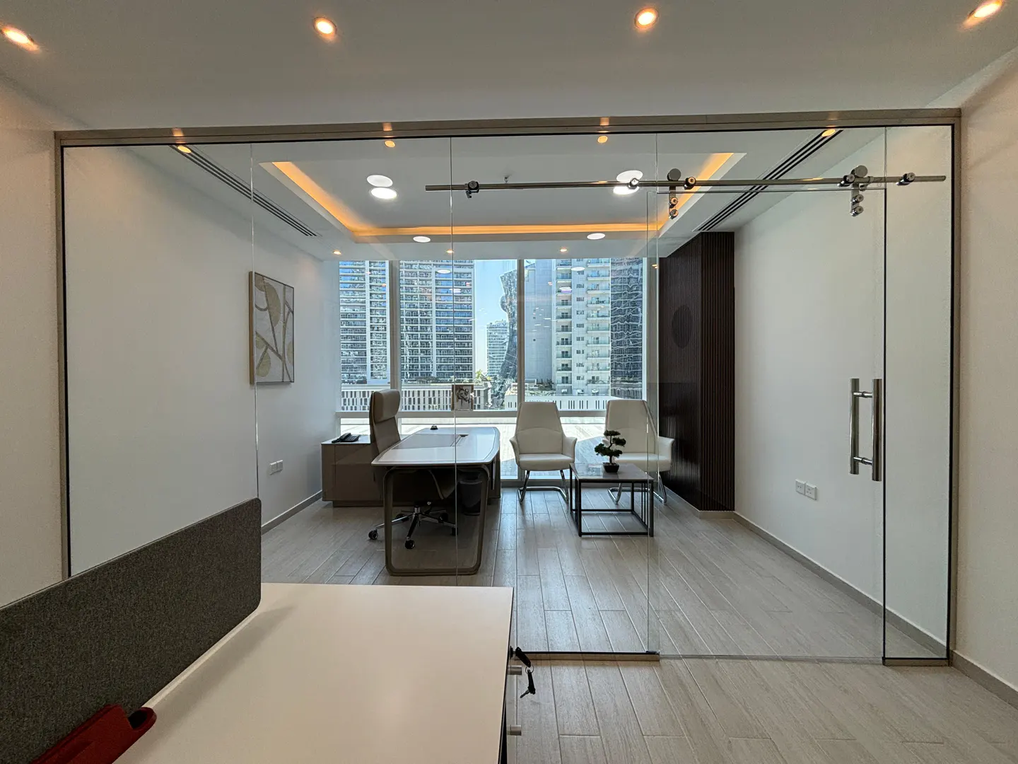 Office interior with glass sliding door. A desk, chairs, and city view are visible through the glass. The walls are white, and the floor is light wood.