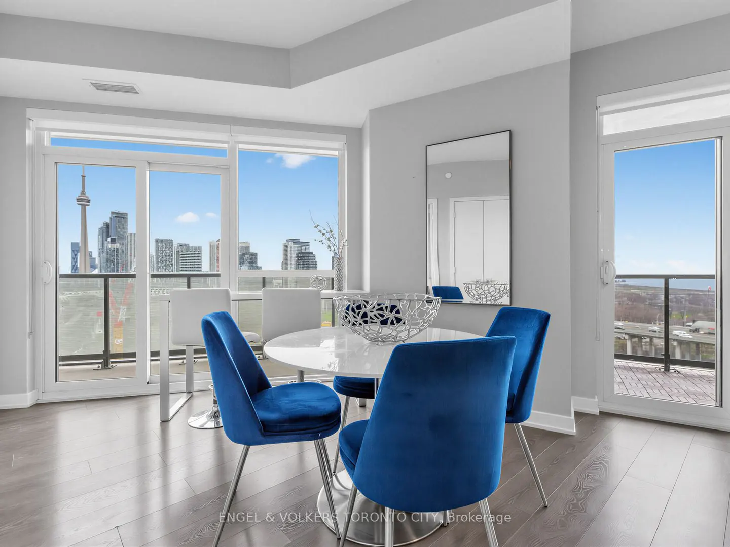 Modern dining area with a white round table, four blue velvet chairs, and a view of the Toronto skyline through windows.