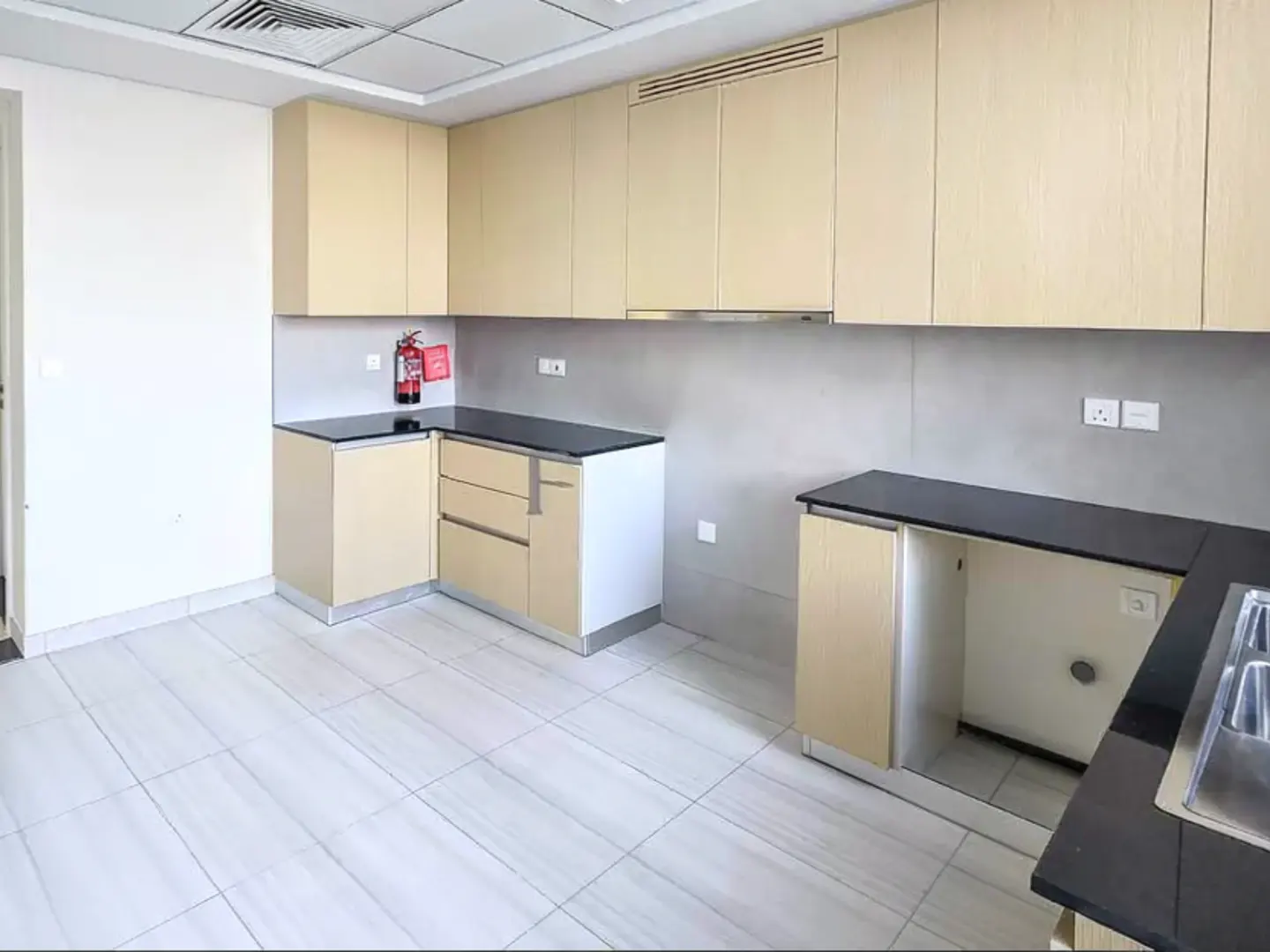 Bright kitchen with light wood cabinets, black countertops, and gray backsplash. A red fire extinguisher is mounted on the wall.
