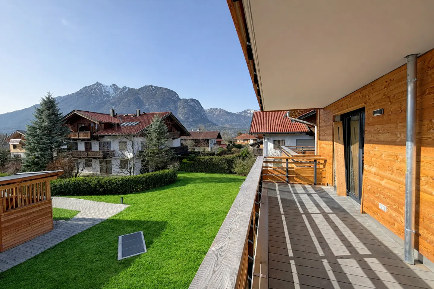 View from a wood balcony overlooking a green lawn, houses, and mountains under a blue sky.