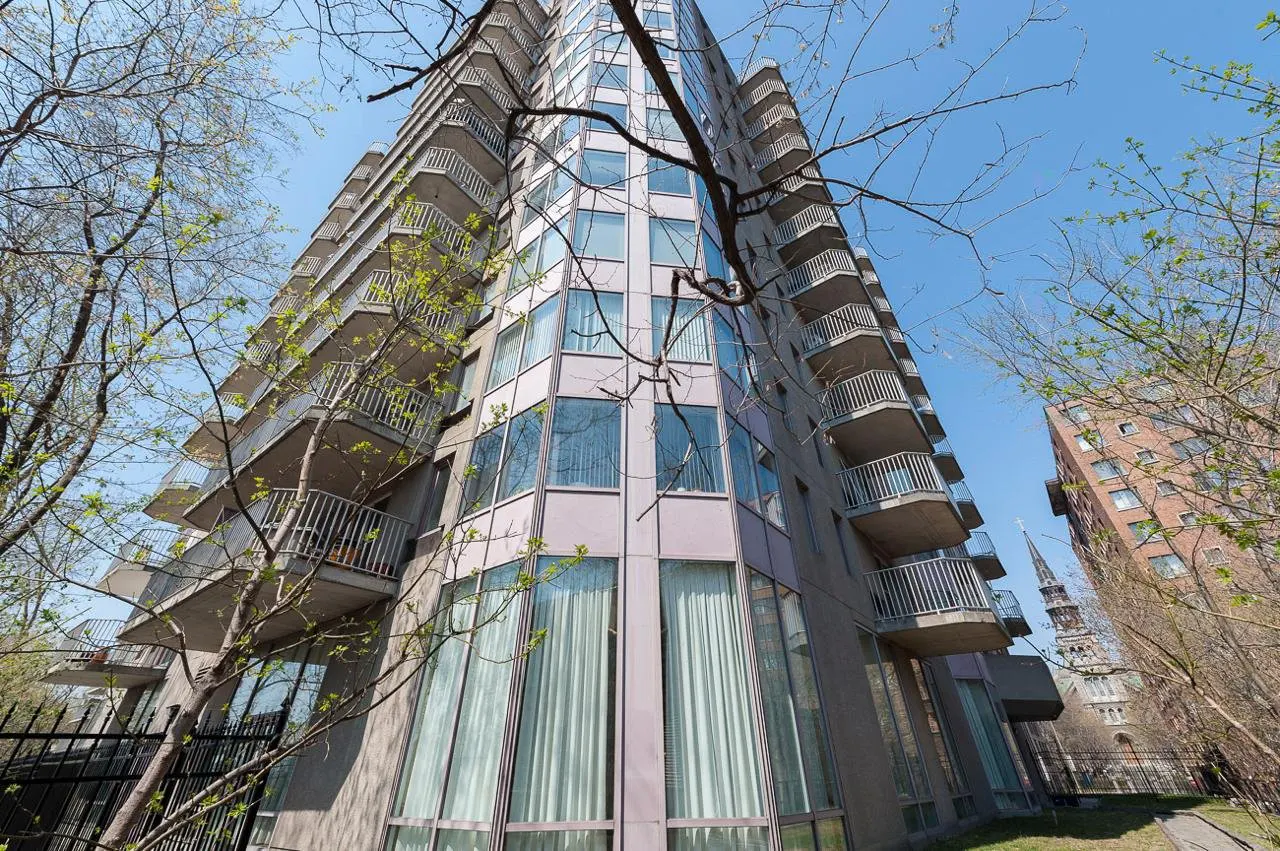 Low-angle view of a tall, gray apartment building with balconies and large windows, framed by trees against a blue sky.