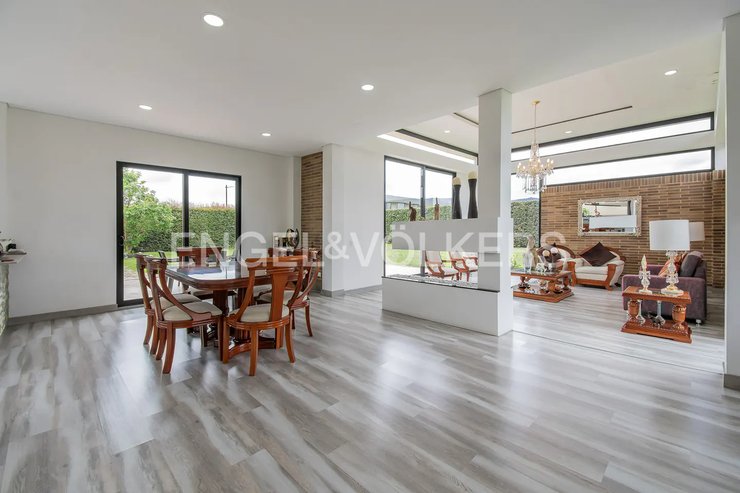 Open-concept home interior with a dining table, living room, and gray wood floors. Large windows offer views of the green outdoors.