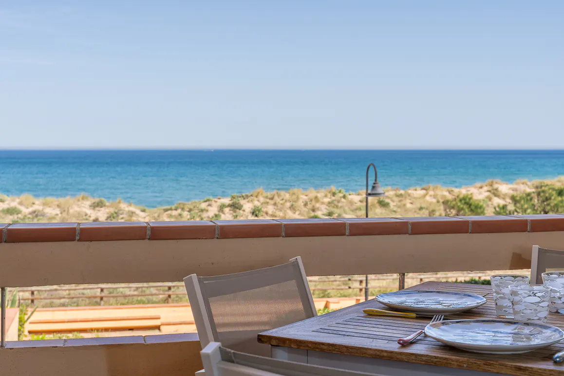 Balcony view of the ocean with a set table. Plates, glasses, and cutlery are on a wooden table with white chairs.