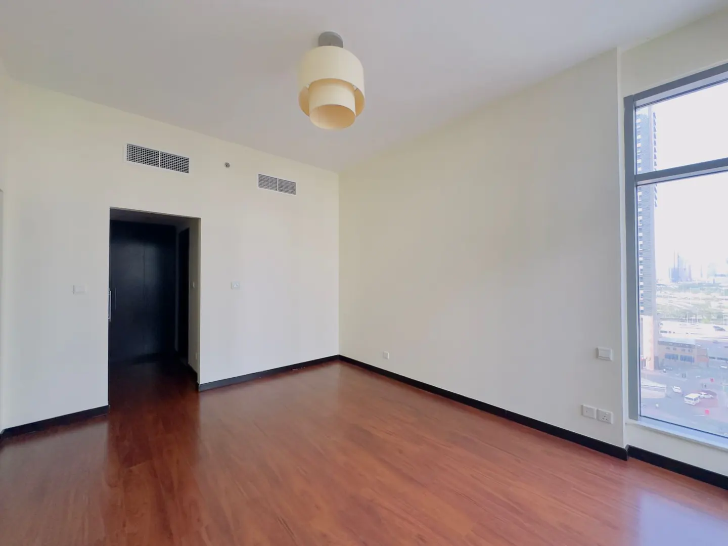Empty room with wood floors, white walls, and a modern light fixture. A large window shows a city view.