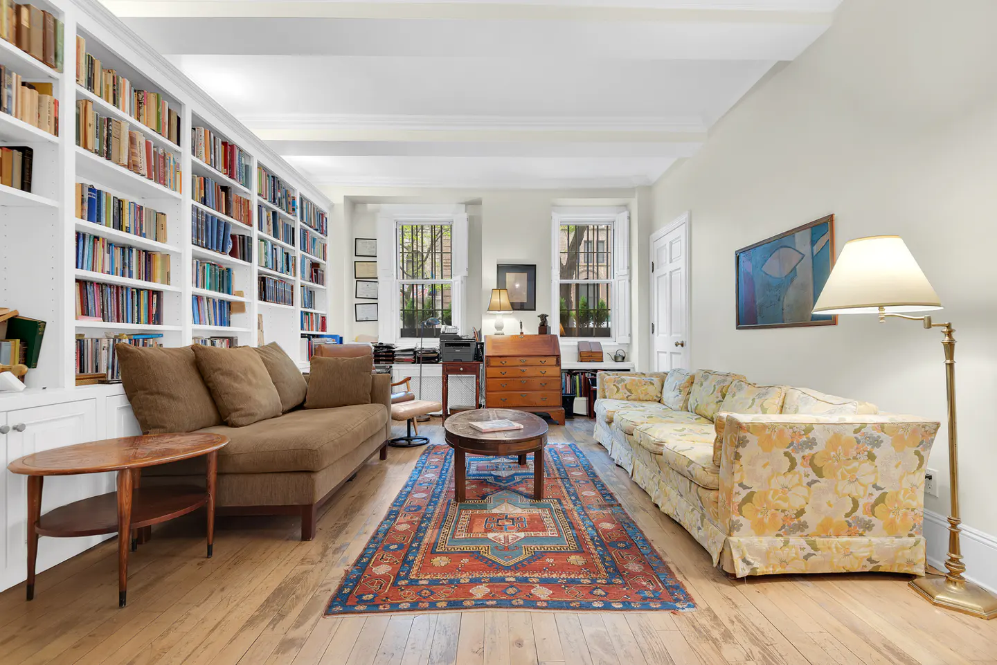 Living room with hardwood floors, a brown sofa, a floral sofa, and a large bookshelf filled with books. A colorful rug is centered in the room.